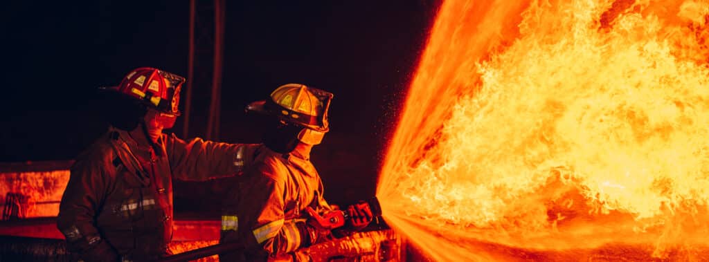Firefighters spraying water and using extinguishers to control the fierce flames, surrounded by smoke that carries asbestos