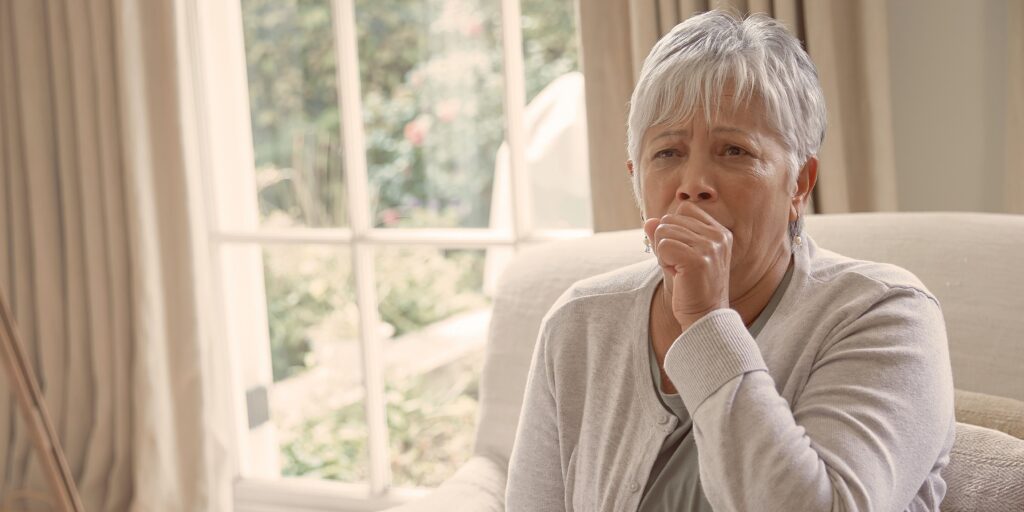 Senior woman with mesothelioma coughing on her couch in her home with a worried expression