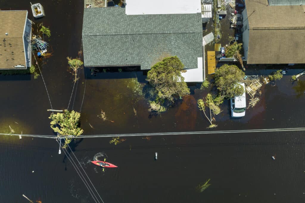 An aerial shot shows a neighborhood street completely flooded, with water reaching the roofs of cars and covering lawns up to the houses. A red kayak with two people can be seen paddling through the water.