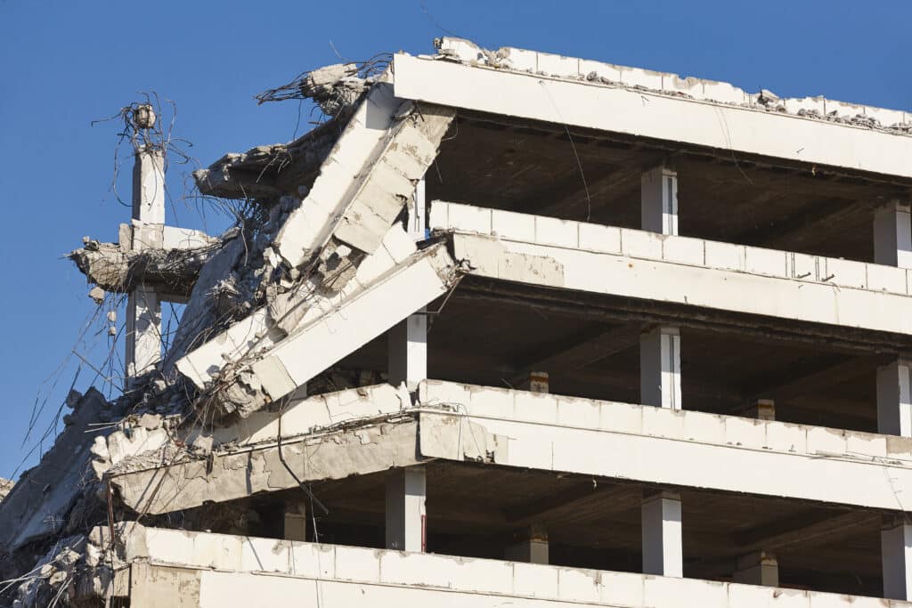 A partially demolished building shows its gutted floors, exposed rebar, and fractured support beams. The remaining structure stands against a clear blue sky.