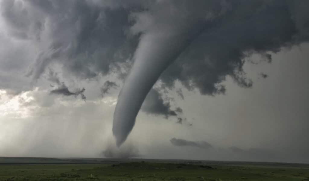 A massive tornado touches down in a rural setting. The powerful funnel cloud extends from a dark, turbulent sky down to a green field, causing dust and debris to swirl at ground level.