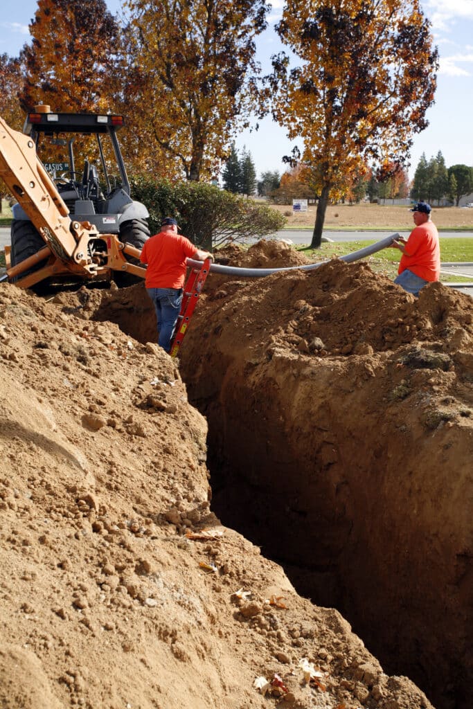 Two men are working together in a construction zone, installing a gray pipe between a deep trench and a tractor.