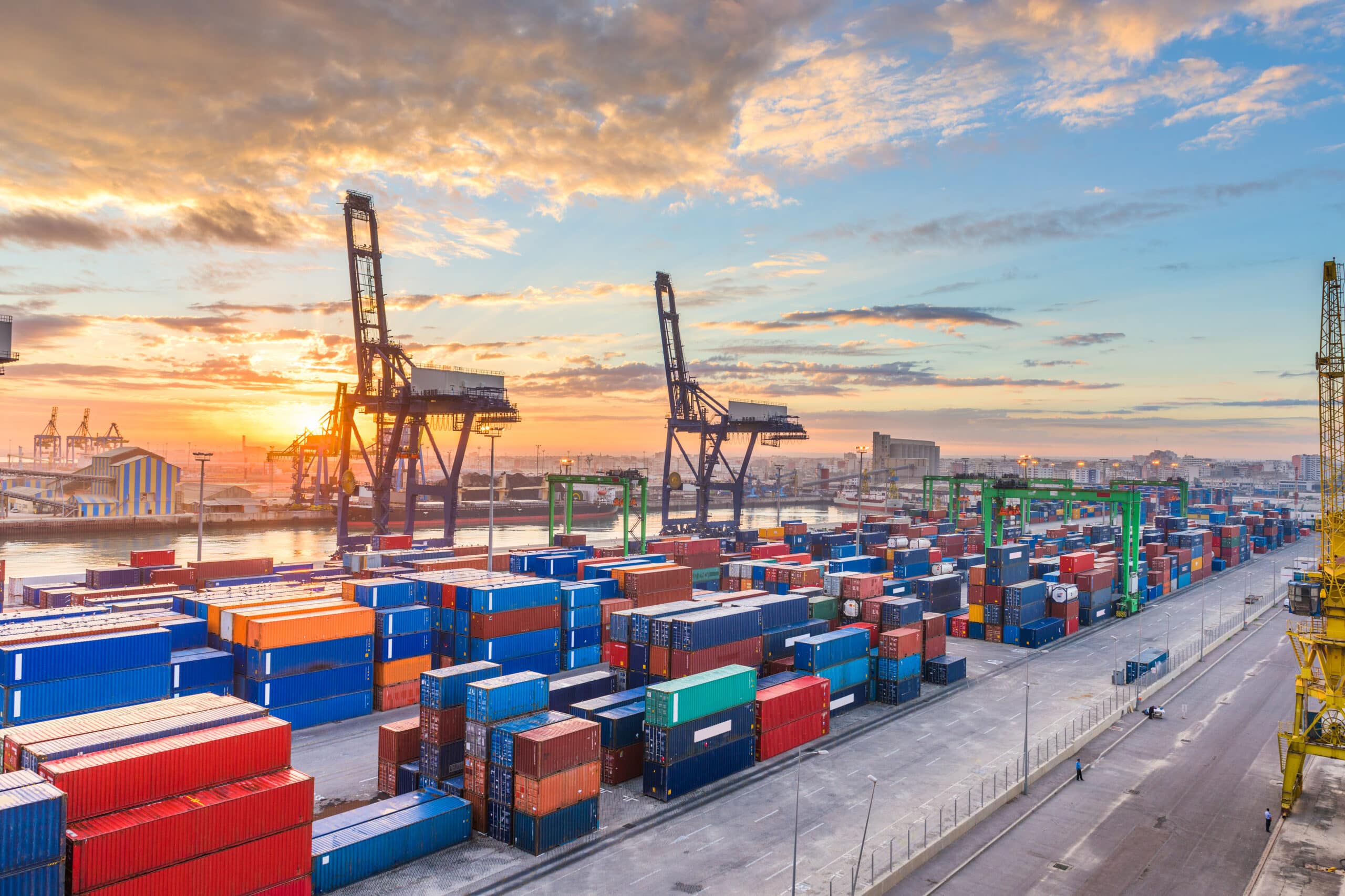 Shipping containers fill a bustling port at sunrise or sunset. Cranes stand silhouetted against the colorful sky, and stacked containers of various colors are positioned around the harbor.