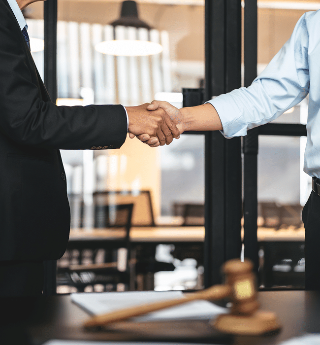 Two business professionals shaking hands across a desk with a judge's gavel and legal documents in the foreground.